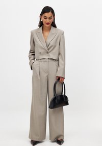 Blazer beige cintré avec double boutons, pantalon beige à jambe large et sac à main noir incurvé. Tissu lisse avec un design sur mesure.