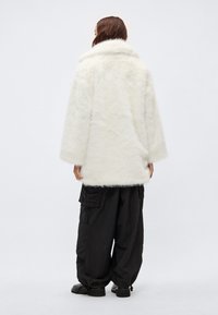 Λευκό παλτό από συνθετική γούνα με φαρδύ γιακά και άνετη γραμμή, φορεμένο πάνω από μαύρο παντελόνι cargo. Το παλτό έχει απαλή υφή και μήκος μέχρι τη μέση.