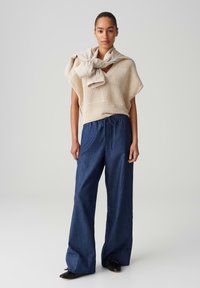 Beige knitted top with a V-neck, paired with navy wide-leg denim pants. A beige lightweight sweater draped over the shoulders. Black shoes.