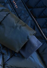 Marineblauer, gesteppter Pufferjackenärmel mit einer glatten, glänzenden Textur. Verfügt über einen kontrastierenden blauen Saum, der mit "BAKER" in dunkler Schrift geprägt ist.