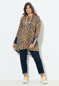 Robe sweat ample bleu marine avec imprimé léopard jaune, cordon de serrage et manches longues ; associée à un jean foncé et des baskets blanches avec des accents jaunes.