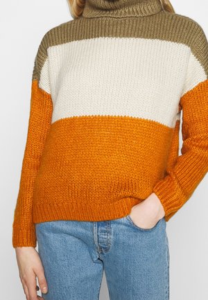 Femme portant un pull à col roulé en tricot avec des rayures horizontales marron, blanches et orange, assorti à un jean bleu.