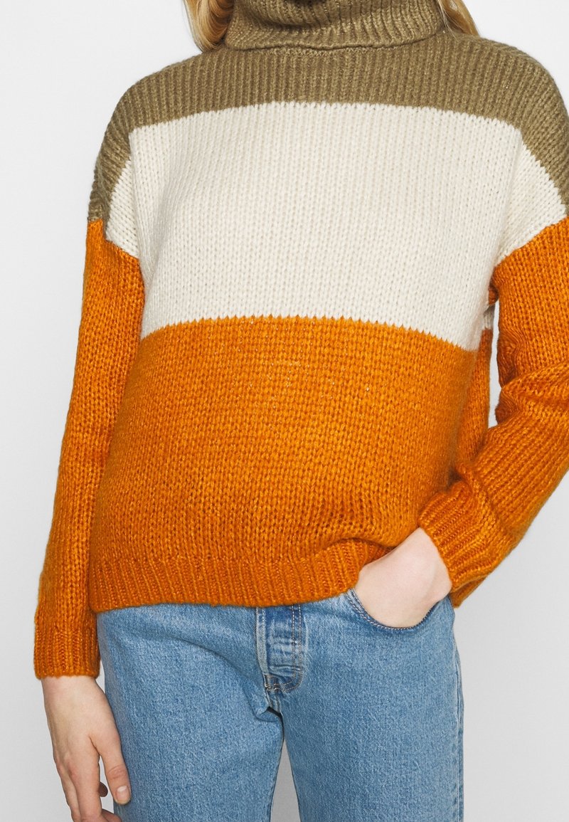 Femme portant un pull à col roulé en tricot avec des rayures horizontales marron, blanches et orange, assorti à un jean bleu.