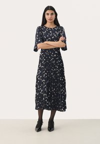 Abito blu navy con pieghe e maniche corte, caratterizzato da un motivo astratto bianco e oro. Indossato con tacchi neri, su uno sfondo neutro.