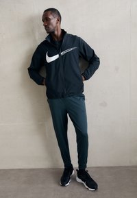 Sort Nike-jakke med et hvidt logo, grønne træningsbukser og sorte sneakers. Glat stof, lynlåslukning og afslappet design.