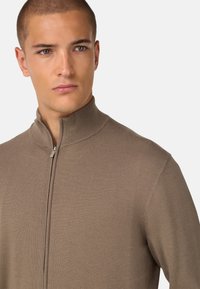 Pruun, tõmblukuga kampsun kõrge kaelusega. Pehme, soonikuline tekstuur ja lähedane disain. Omab peent sära ja minimalismi stiili.