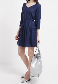 Abito blu navy semi-attillato con scollatura a V, maniche 3/4 e un cordoncino regolabile in vita. Abbinato a una borsa a tracolla metallica argento.
