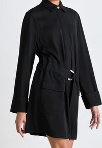 Donna in un cappotto nero lungo fino al ginocchio, con cintura, grandi polsini, tasche applicate e colletto con bottoni, su uno sfondo semplice.