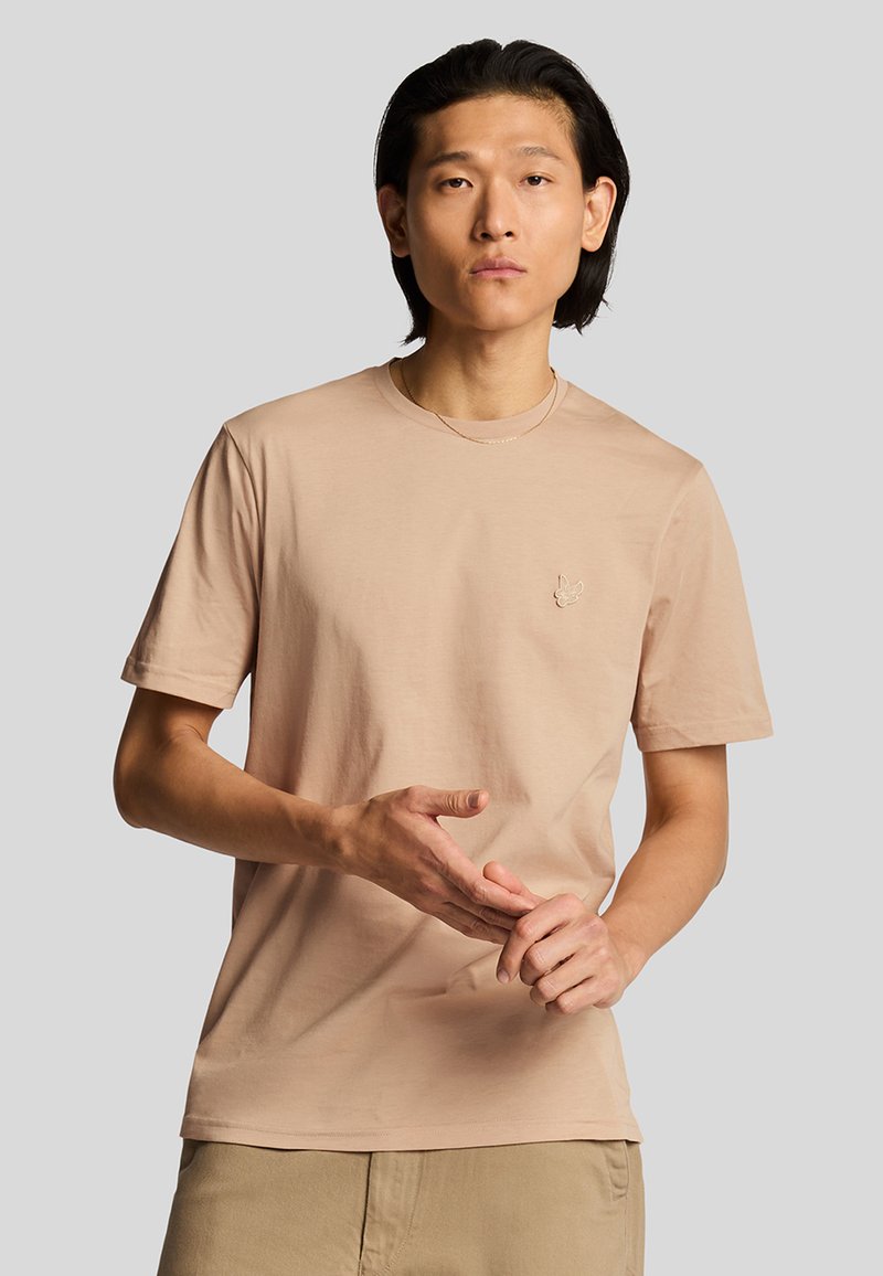 T-shirt di cotone beige chiaro con maniche corte e colletto tondo, con un piccolo logo ricamato sul petto. Tessuto liscio.