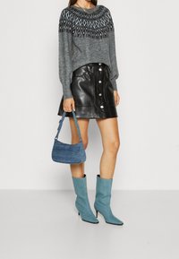 Femme portant un pull gris à motifs, une jupe en cuir noir à boutons, des bottes à talons bleu clair mi-mollet, et tenant un sac à main texturé bleu.