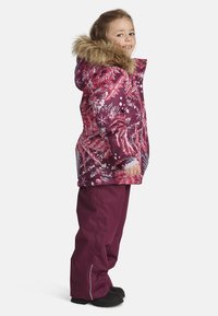 Paars en roze winterjack met een met bont gevoerde capuchon, voorzien van een bladpatroon. Gecombineerd met effen paarse broek en zwarte laarzen.