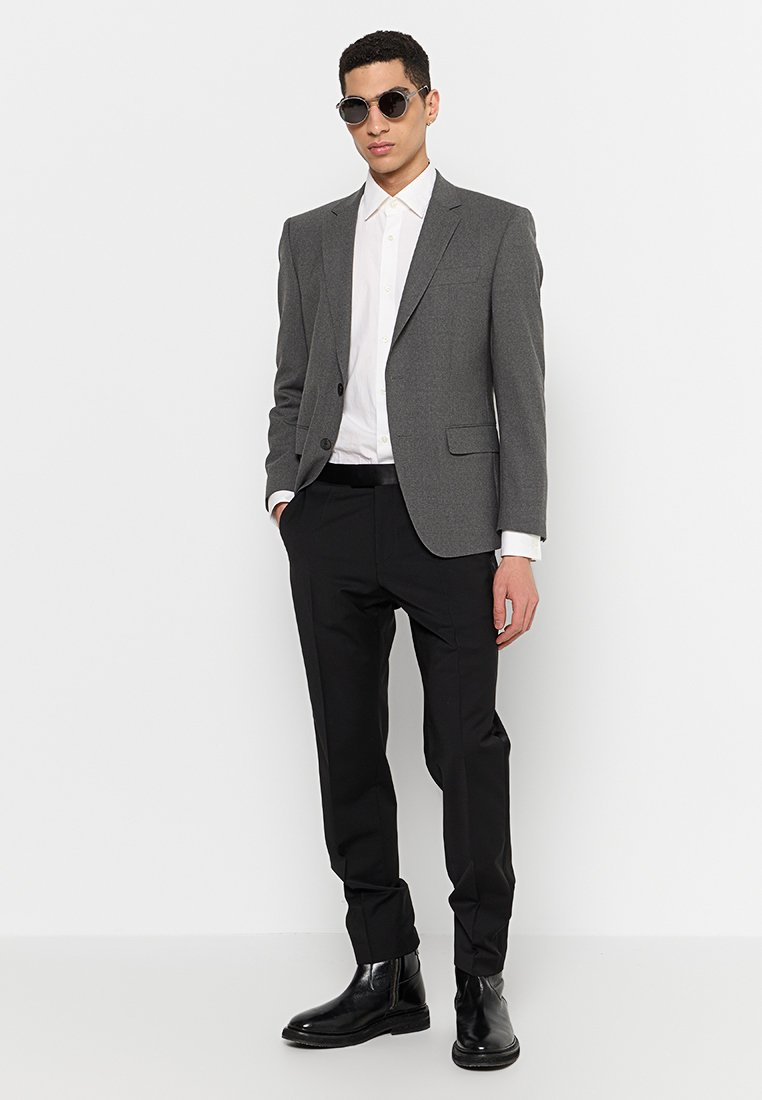 Blazer gris sur une chemise blanche, pantalon noir et bottes noires. Le blazer a un col cranté et deux poches avant.
