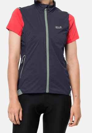 Personne portant un gilet zippé bleu marine sur un t-shirt rouge à manches courtes et un cuissard de cyclisme noir rembourré, debout les bras détendus.