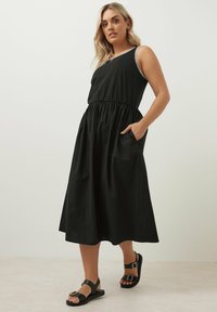 Svart ärmlös klänning med en åtdragen midja och elastiskt band, med sidofickor och kontrasterande kantning. Kombinerad med svarta sandaler.