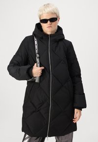 Crna puffer jakna s dijamantnim kvadratnim dizajnom, sa zatvaračem sprijeda i kapuljačom. Nošena preko metalik srebrnih hlača, s crnom torbom na remenu.