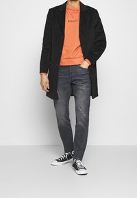 Černý kabát ze směsi vlny, oranžová mikina s potiskem "MENACE", oprané tmavé džíny a černé vysoké tenisky s bílou podrážkou.