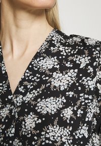 Haut noir à motifs floraux avec un col en V, décoré de motifs floraux bleu clair et beige. Le tissu est lisse avec un léger éclat.