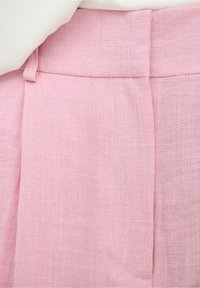 Pantalon en lin rose clair avec une ceinture devant et des passants pour ceinture, porté avec un haut blanc rentré à l’intérieur.