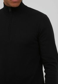 Schwarzer, langärmeliger Pullover mit hohem Kragen und halbem Reißverschluss. Hergestellt aus einem glatten, dehnbaren Stoff mit einer taillierten Silhouette.