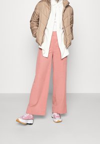 Beżowa pikowana kurtka, biała koszula z guzikami i lekkie różowe spodnie o szerokich nogawkach. Różowe sneakersy z białymi i czarnymi detalami.