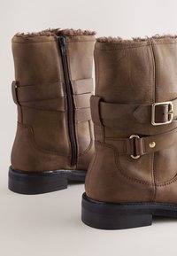 Bottes en cuir marron avec une fermeture éclair latérale, des accents de sangle avec une boucle dorée, une doublure en fourrure marron moelleuse et une semelle noire robuste.