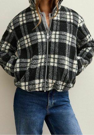 Personne portant une veste polaire à carreaux noir et blanc avec une fermeture éclair à l'avant et un jean bleu, les mains dans les poches de la veste.