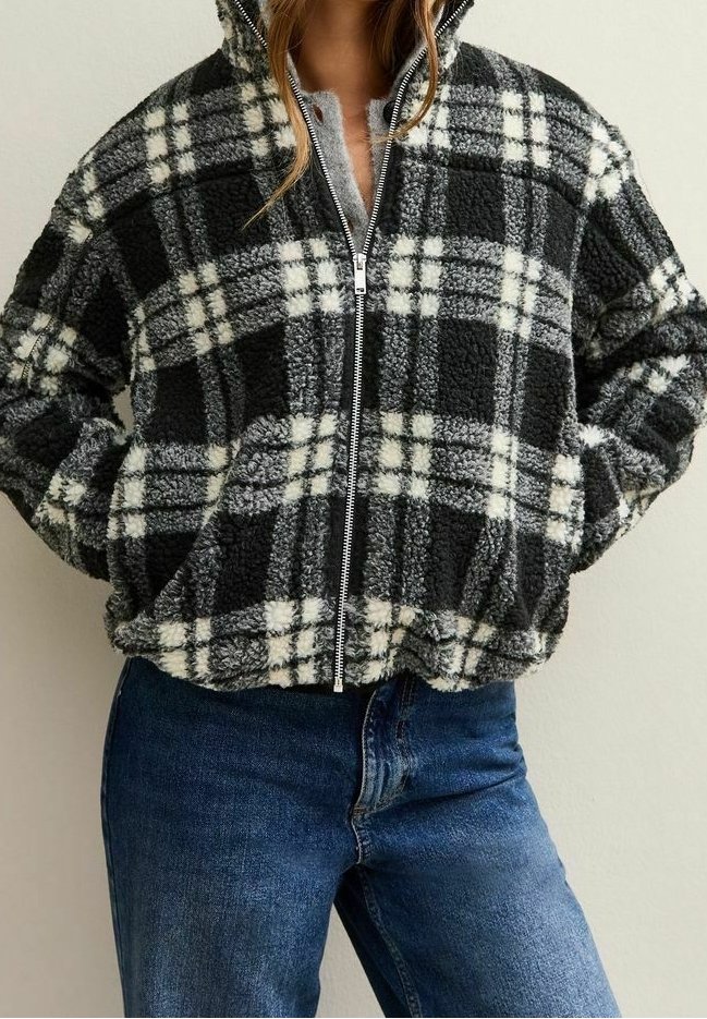 Personne portant une veste polaire à carreaux noir et blanc avec une fermeture éclair à l'avant et un jean bleu, les mains dans les poches de la veste.