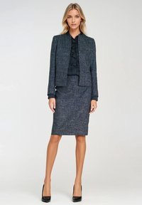 Kobieta ubrana w ciemnoszary garnitur z tweedu z falbaniastą czarną bluzką i czarnymi szpilkami, stojąca na tle gładkiego białego tła.