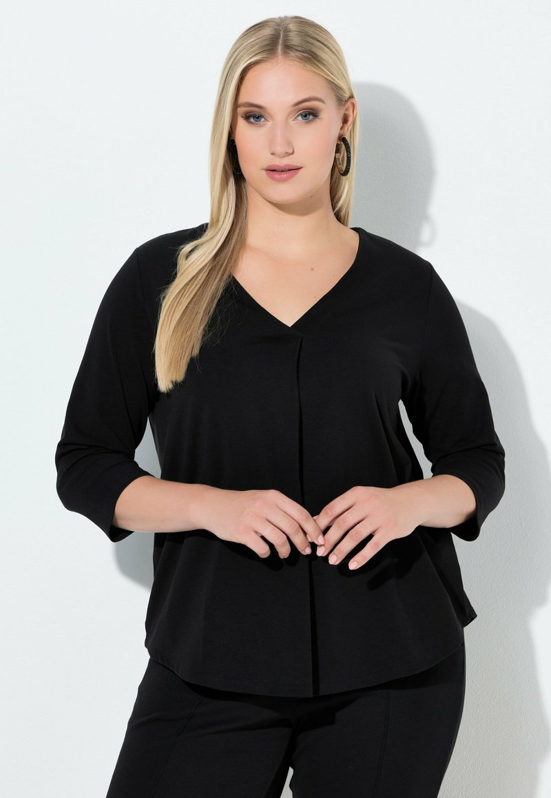 Schwarzes Langarmshirt mit V-Ausschnitt und dezentem Drapée vorne. Glattes, weiches Material mit lockerem Sitz, kombiniert mit schwarzen Hosen.