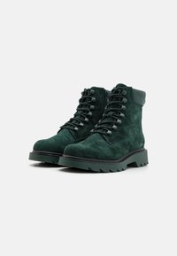 Des bottes chevilles en suede vert présentent des œillets en métal, des lacets épais, une semelle en caoutchouc épaisse et un accent noir lisse au niveau du talon.