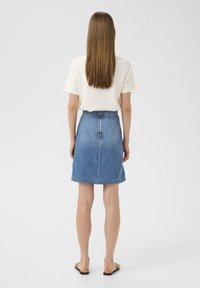 Jupe en denim avec une légère forme évasée, de couleur bleu moyen, fermeture éclair au dos et ourlet cousu. Associée à un haut blanc à manches courtes.