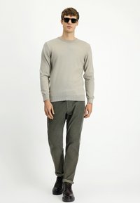 Mężczyzna w beżowym swetrze z długim rękawem, ciemnozielonych spodniach, czarnych butach eleganckich i czarnych okularach przeciwsłonecznych idzie na tle jednolitego białego tła.