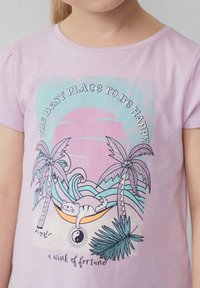 Lila kurzärmliges T-Shirt mit einem grafischen Design, das Palmen, eine sich ausruhende Katze, Wellen und den Text "Der beste Ort, um glücklich zu sein" zeigt.