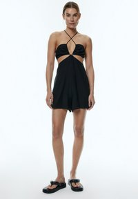 Schwarzer Cut-Out-Jumpsuit mit Neckholder, überkreuzten Trägern und lockerem Schnitt. Kombiniert mit schwarzen klobigen Flip-Flops. Glatte Stofftextur.