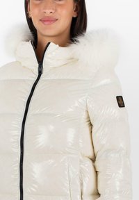 Giacca imbottita realizzata in materiale lucido di colore crema, con cappuccio in pelliccia sintetica, zip nera e patch con logo sulla manica.