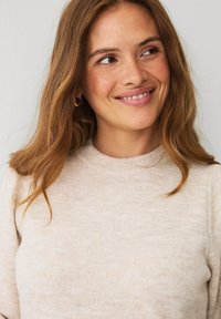 Kvinde med langt lysbrunt hår, iført små hoop øreringe og en beige sweater, der smiler og ser til højre mod en ensfarvet baggrund.