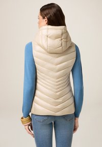 Beige gepolsterte Weste mit Chevron-Design, hohem Kragen und Kapuze, kombiniert mit einem blauen Langarmtop. Glatte Textur und taillierter Schnitt.