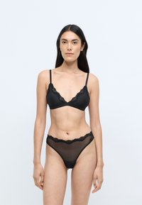 Brassière en dentelle noire avec des bonnets en triangle et des bretelles réglables, associée à un string en dentelle noire transparente assorti. Bords délicatement festonnés.