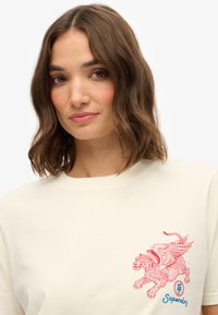 Creme crewneck t-shirt lavet af blødt bomuld med et rødt vingerløve-grafik på venstre bryst og en blå "Superdry" logo under det.