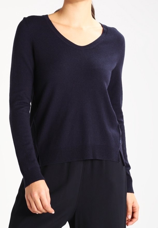 Woman wearing a long-sleeve, dark navy V-neck sweater and dark pants with hands relaxed by her sides.