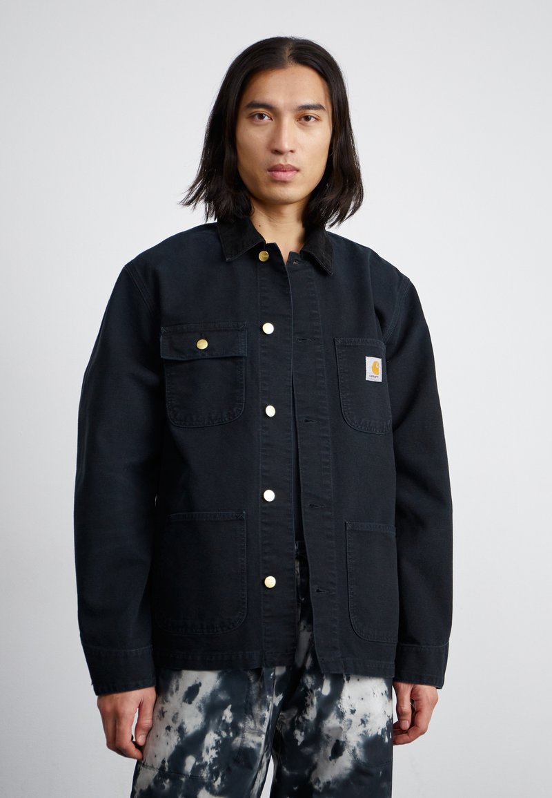 Schwarze Jeansjacke mit Kragen, goldenen Druckknöpfen und zwei Fronttaschen; verfügt über ein kleines Logo-Patch auf der linken Brust.