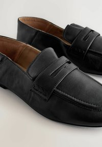 Black leather loafers featuring a smooth texture, rounded toe, and a decorative strap with vertical slits. Lined interior in tan.