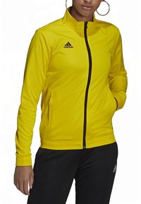 Kvinde iført en gul Adidas zip-up jakke med sort lynlås og sorte Adidas bukser, hænderne i lommerne, hoops-øreringe synlige.