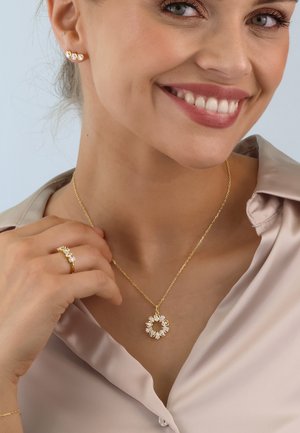 Goldene Anhängerhalskette mit kreisförmigem Design, verziert mit klaren Steinen; passender goldener Ring und Ohrringe mit ähnlichen Steinakzenten.