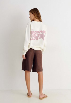 Femme debout à l'intérieur portant un sweat blanc avec le texte rose « Brownie Girls Club » et un short marron jusqu'aux genoux ainsi que des sandales.