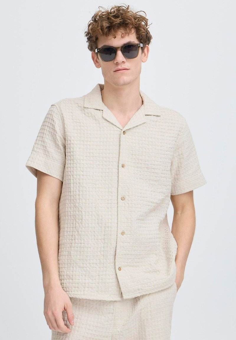 Camisa corta de manga corta color beige claro con un patrón texturizado, que presenta un cuello y tres botones. Llevada con gafas de sol negras.