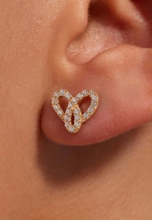 Gold-tone heart-shaped earring adorned with clear rhinestones, featuring a smooth surface and intertwined design. Applied on skin background.