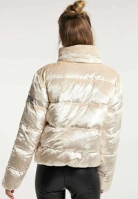 Chaqueta acolchada de terciopelo beige claro con un cuello alto, que presenta un diseño acolchado y un corte ajustado. Detalle del logo negro en la manga.