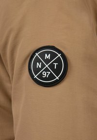 Hnedá tkanina s jemnou textúrou, s kruhovou čiernou nášivkou označenou "NMT 97" s bielymi značkami, umiestnenou na hornej časti.