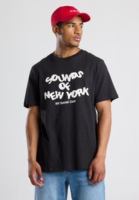 Black cotton t-shirt features white "SOUNDS OF NEW YORK" text. Paired with a red baseball cap. Simple design, casual fit.